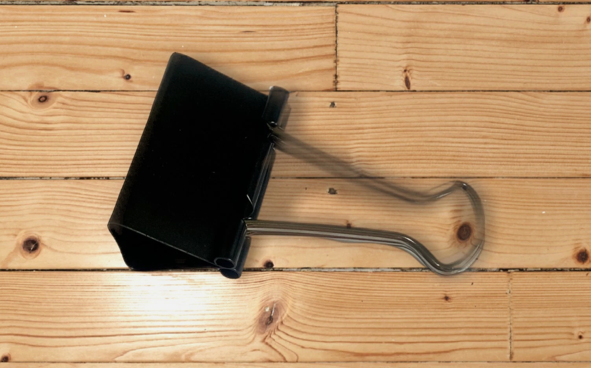A black binder clip with a clear plastic inner band, placed on a light-colored wooden plank surface.