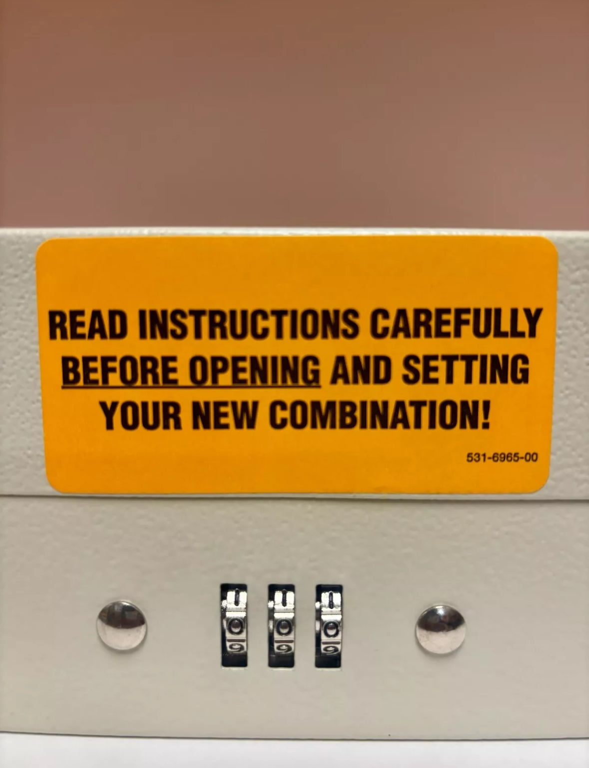 A close-up of a beige security box or safe with a bright yellow warning label that reads, 'READ INSTRUCTIONS CAREFULLY BEFORE OPENING AND SETTING YOUR NEW COMBINATION!' above three black combination dials.