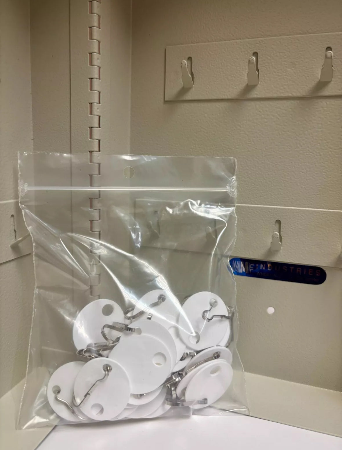 A clear plastic zip-top bag containing multiple white round adhesive labels with metal hanging loops, placed on a beige shelf with hooks in the background.