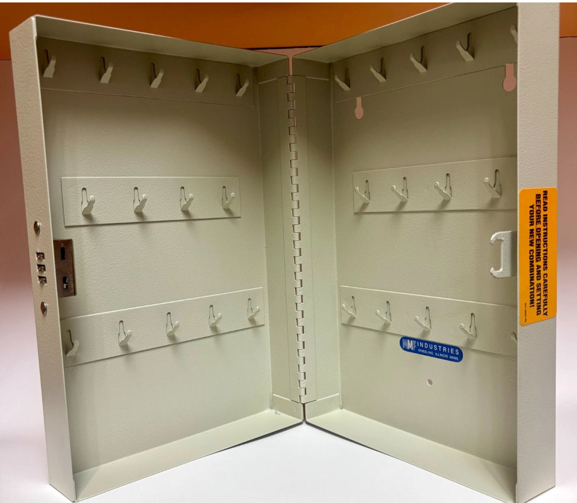 An open metal wall-mounted key box or storage cabinet with multiple hooks on the interior panels, photographed against a plain light background with an orange backdrop visible at the top.